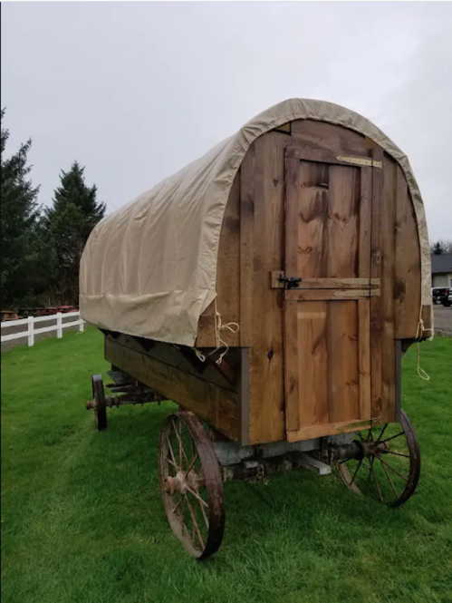 A wooden covered wagon on wheels, set on green grass with a cloudy sky in the background.