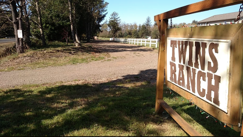 Sign for "Twins Ranch" with a gravel path and trees in the background on a sunny day.