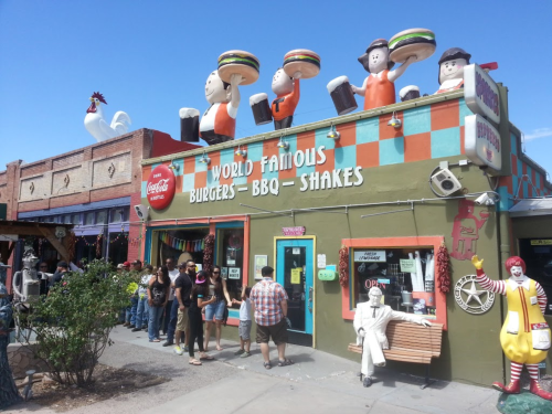 A colorful restaurant with burger and shake signs, featuring cartoon figures on the roof and a line of customers outside.