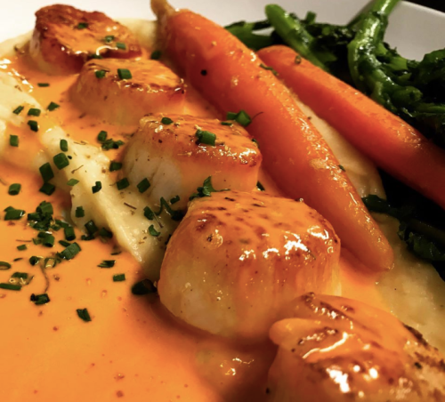A close-up of seared scallops on a bed of sauce, accompanied by carrots and green vegetables.