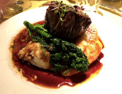 A gourmet dish featuring a tender piece of meat atop creamy mashed potatoes and sautéed greens, drizzled with sauce.