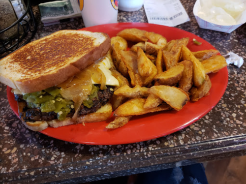 A plate with a grilled sandwich topped with cheese, pickles, and a side of golden-brown potato wedges.