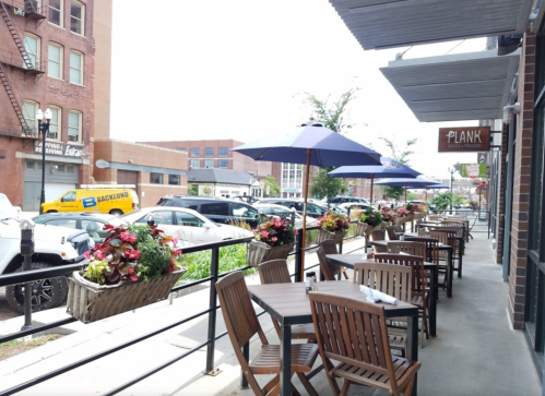 Outdoor dining area with wooden tables, umbrellas, and flower planters along a sidewalk in a city setting.
