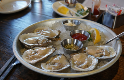 A platter of fresh oysters on ice, served with lemon wedges and dipping sauces in small metal bowls.