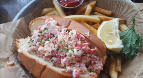 A lobster roll filled with lobster meat, served with fries, a lemon wedge, and a small cup of ketchup.
