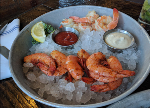 A metal platter filled with ice, featuring shrimp, lemon, and two dipping sauces.