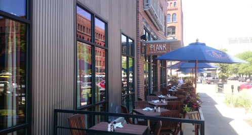 Outdoor dining area of a restaurant named "Plank," featuring tables, umbrellas, and a sunny urban setting.