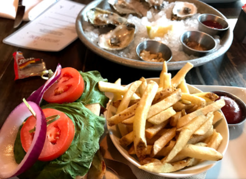 A plate with a sandwich topped with lettuce, tomato, and onion, alongside a serving of fries and a platter of oysters.