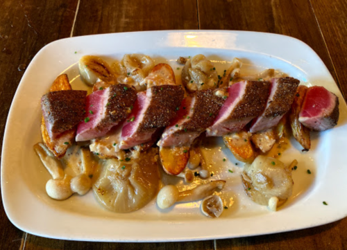 A plate of sliced seared meat on a bed of sautéed vegetables and creamy sauce, garnished with herbs.