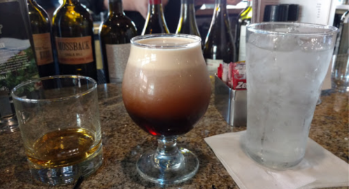 A bar scene featuring a layered drink in a glass, a whiskey glass, and a tall glass of water on a granite countertop.