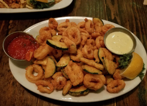 A plate of fried calamari with zucchini slices, served with marinara and a creamy dipping sauce, garnished with lemon.