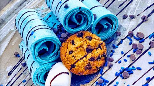 Blue rolled ice cream with chocolate drizzle, a chocolate chip cookie, and a marshmallow, scattered with chocolate chips.