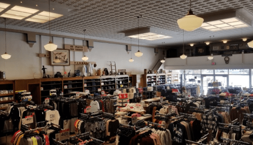A spacious retail store filled with clothing racks, shelves, and vintage decor, featuring large windows and bright lighting.