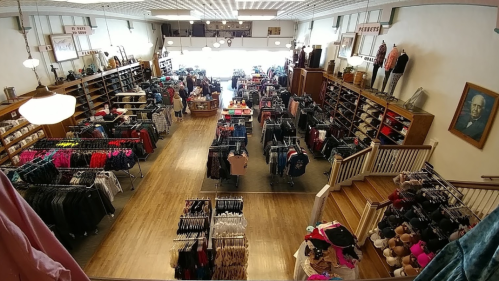 A spacious thrift store filled with clothing racks, shelves, and shoppers, featuring a bright, welcoming atmosphere.
