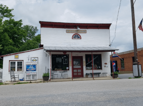 A quaint general store with a white exterior, red accents, and a porch, surrounded by greenery and a nearby brick building.