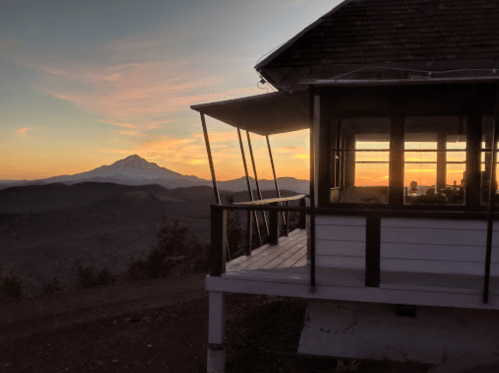 A cabin porch overlooks a mountain at sunset, with warm colors in the sky and a silhouette of the landscape.