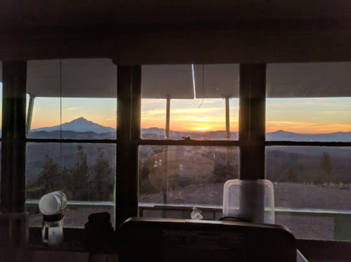Sunset view from a cabin window, showcasing mountains and a colorful sky, with trees in the foreground.