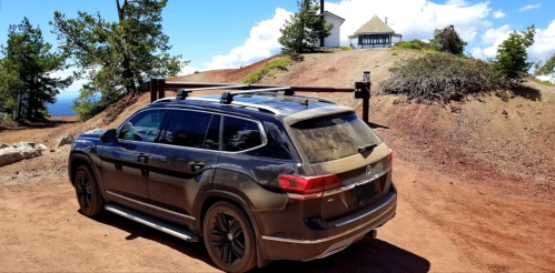 A dark SUV parked on a dirt road near a small building and trees under a blue sky with clouds.