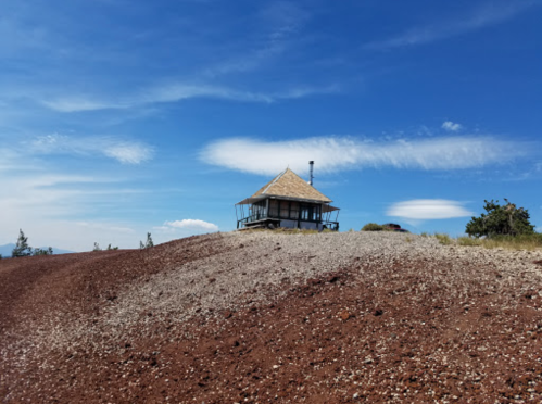 A small wooden house on a hill with a gravel path, surrounded by blue sky and scattered clouds.