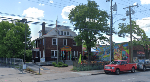 A colorful restaurant with outdoor seating, surrounded by greenery and a vibrant mural, set in a brick building.