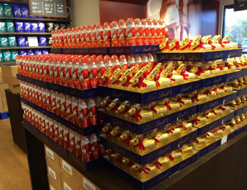 A display of stacked chocolate boxes, featuring red and gold Lindt chocolates, in a store setting.