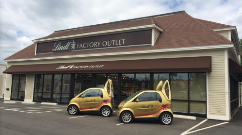 Lindt Factory Outlet storefront with two gold Smart cars parked in front, featuring chocolate branding.
