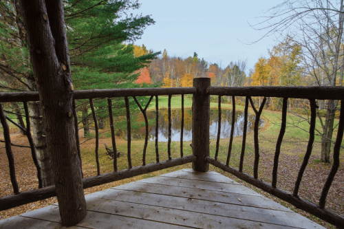 View from a wooden balcony overlooking a serene pond surrounded by autumn foliage and trees.