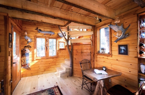 Cozy wooden interior of a cabin with a spiral staircase, rustic decor, and a small table by the window.