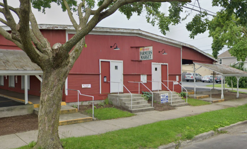 Red barn-style building with a sign reading "Farmers Market," featuring steps and a grassy area in front.