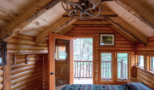 Cozy wooden cabin interior with a sloped ceiling, large windows, and rustic decor, overlooking a forested area.