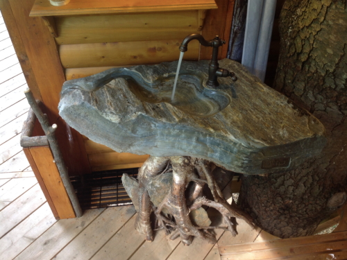 A unique sink made from a large stone slab with a tree root base and a vintage faucet, set in a wooden cabin.