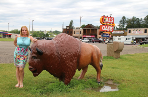 A woman stands next to a large bison statue outside Al's Oasis, with shops and trees in the background.