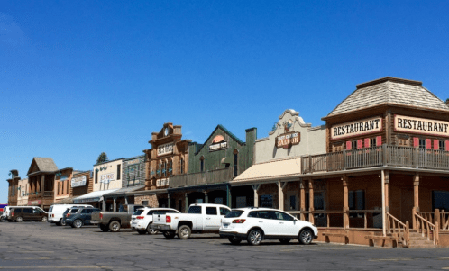 A row of Western-style buildings with shops and restaurants, parked cars in front, under a clear blue sky.