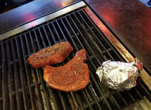 Two seasoned meat cuts grilling on a barbecue, alongside a foil-wrapped item.
