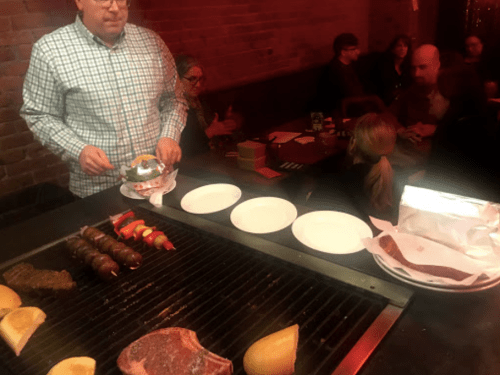 A chef serves food at a grill, with skewers and steaks cooking, while diners enjoy their meals in a cozy restaurant.