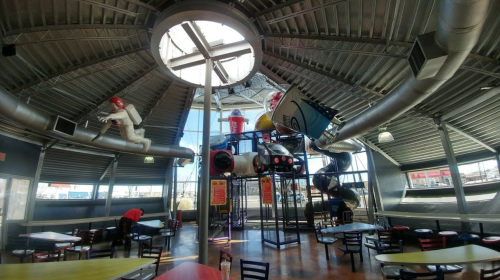 Indoor play area with colorful slides and climbing structures, featuring a circular skylight and tables for dining.