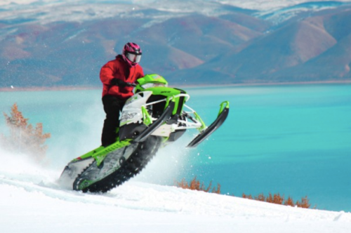 A person in a red jacket rides a green snowmobile over snowy terrain with a turquoise lake in the background.
