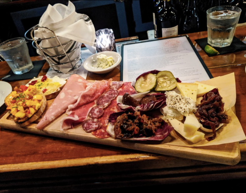 A wooden platter with assorted meats, cheeses, pickles, and garnishes, accompanied by drinks and a menu in the background.