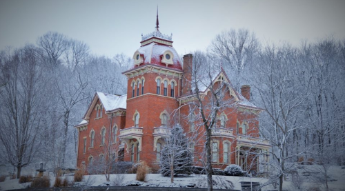 A grand, red brick mansion with a tower, surrounded by snow-covered trees in a winter landscape.