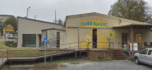 A colorful glass blowing studio with a flower mural, surrounded by gravel and a ramp, on a cloudy day.