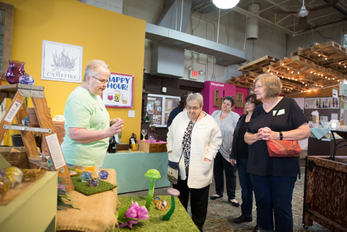 A group of women observes a display of colorful crafts at a market event, with one woman demonstrating an item.