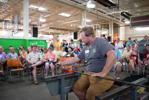 A glassblower demonstrates the art of glassblowing to an audience in a spacious workshop.
