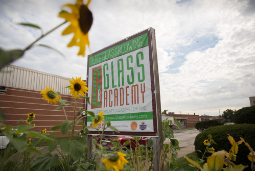 Sign for Glass Academy with sunflowers in the foreground, promoting glassblowing classes and events.