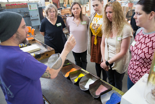 A group of women watches a man demonstrate glass-making techniques, with colorful materials displayed in bowls.