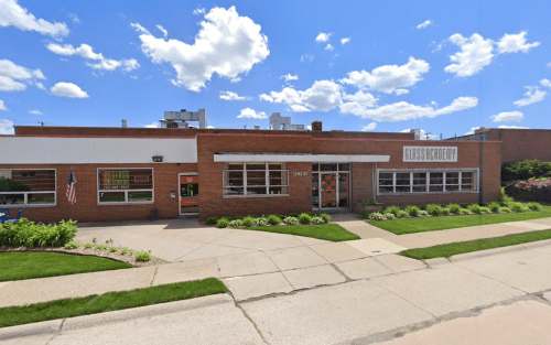 A brick building with large windows, an American flag, and a sign reading "Glass Academy" under a blue sky with clouds.