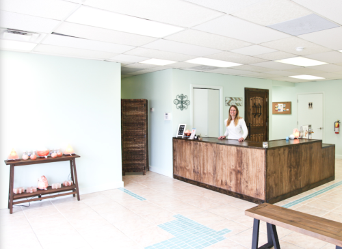 A bright reception area with a wooden desk, a smiling woman, and decorative elements in a light-colored room.