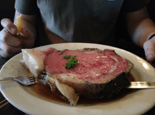 A large, juicy prime rib steak on a plate, garnished with parsley, with a fork and knife beside it.