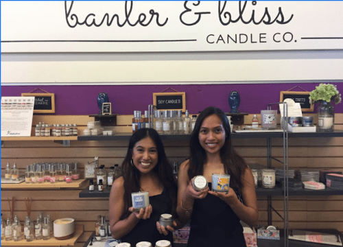 Two women hold candles in front of a display of various candle products at Banter & Bliss Candle Co.