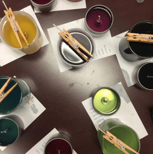 A flat lay of colorful candle jars with wooden clothespins, arranged on a table with paper labels underneath.