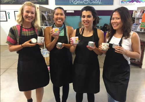 Four women in black aprons hold small containers, smiling in a colorful craft shop.